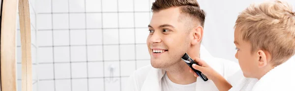 Son holding electric razor and shaving face of father smiling and looking at mirror, banner — Stock Photo