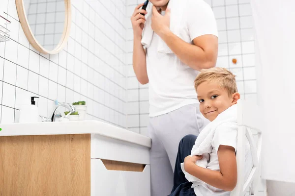 Positive boy looking at camera while sitting on chair near man shaving with electric razor in bathroom — Stock Photo