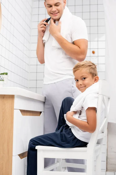 Sorridente figlio guardando la fotocamera mentre seduto sulla sedia vicino felice padre rasatura con rasoio elettrico in bagno — Foto stock