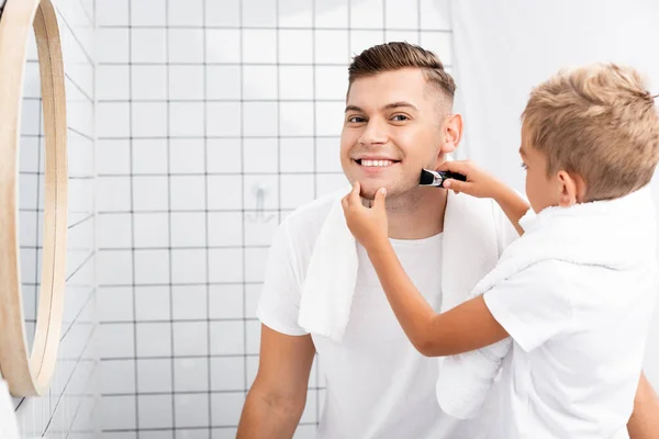 Fils rasage joue de père avec rasoir électrique près du miroir dans la salle de bain — Photo de stock