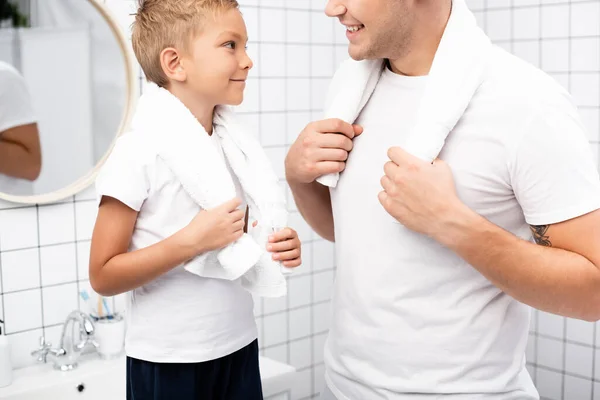 Feliz hijo con toalla en el cuello mirando al padre mientras está cerca del lavabo en el baño - foto de stock