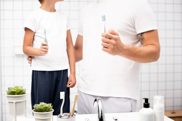 Vue recadrée du père et du fils tenant des brosses à dents près de l'évier avec des articles de toilette dans la salle de bains — Photo de stock