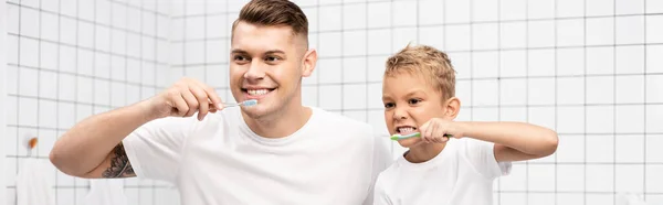 Padre e hijo mostrando dientes mientras sostienen cepillos de dientes en el baño, pancarta - foto de stock