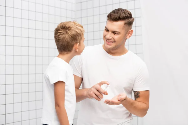 Улыбающийся отец смотрит на сына, сжимая пену для бритья в ванной — стоковое фото