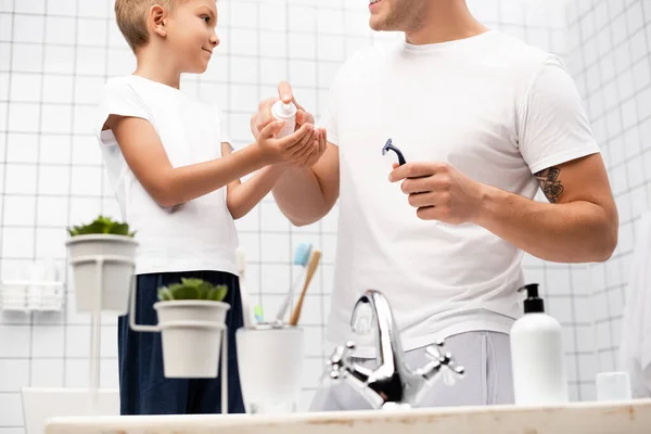 Hijo positivo mirando a padre sosteniendo afeitadora de seguridad mientras se aprieta espuma de afeitar en las manos en el baño en primer plano borrosa - foto de stock