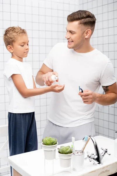 Glücklicher Vater mit Sicherheitsrasierer schaut Sohn an, während er Rasierschaum in der Nähe des Waschbeckens im Badezimmer quetscht — Stockfoto