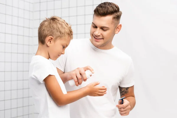 Sonriente padre con afeitadora de seguridad apretando espuma de afeitar en las manos del hijo en el baño - foto de stock