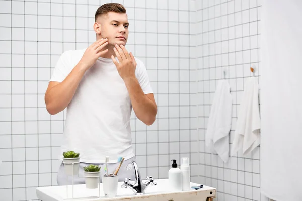 Junger erwachsener Mann berührt Gesicht, während er in der Nähe des Waschbeckens mit Toilettenartikeln im Badezimmer steht — Stockfoto