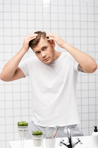 Jeune homme adulte vérifiant les cheveux tout en se tenant près du lavabo dans la salle de bain — Photo de stock