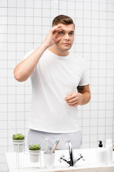 Young adult man touching skin around eye while standing near sink in bathroom — Stock Photo