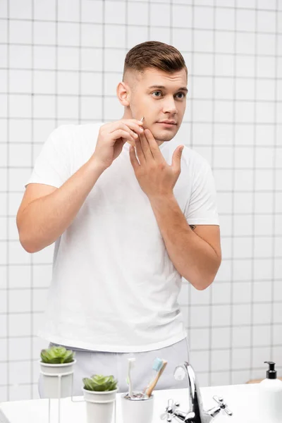 Jeune homme adulte arrachant les cheveux sur la joue avec une pince à épiler dans la salle de bain — Photo de stock