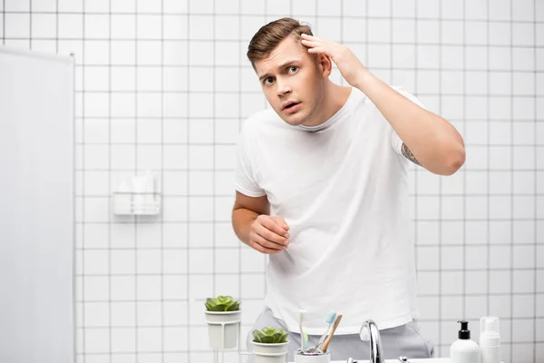 Preoccupato giovane uomo adulto controllare i capelli, mentre in piedi vicino lavandino in bagno — Foto stock