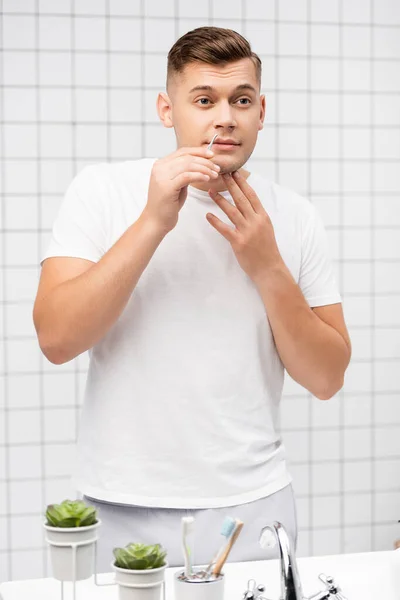 Концентрированный молодой мужчина держит пинцет у губ в ванной комнате — стоковое фото
