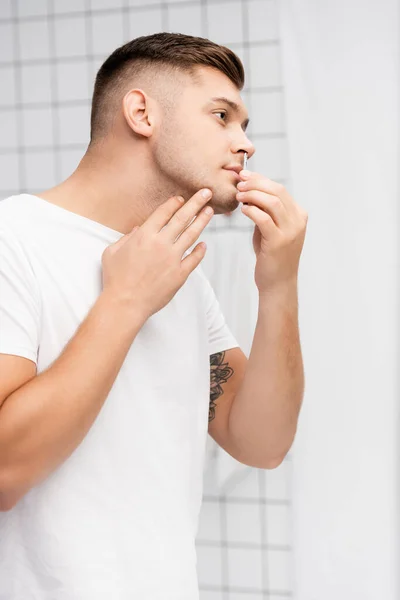 Side view of focused man looking away while plucking hair from nose in bathroom — Stock Photo