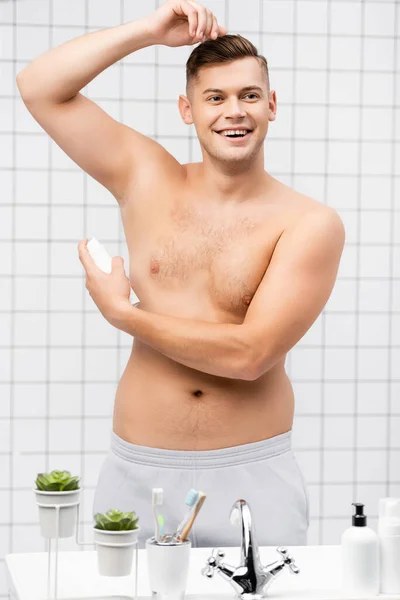 Посміхаючись без сорочки людина використовує дезодорант, стоячи біля раковини з туалетно-косметичними засобами у ванній — стокове фото