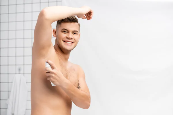 Homem sem camisa feliz olhando para a câmera ao usar desodorizante no banheiro — Fotografia de Stock