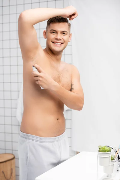 Cheerful shirtless man looking at camera while spraying antiperspirant in bathroom — Stock Photo