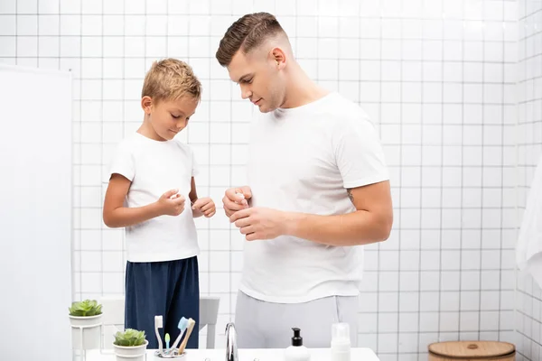 Positive father looking at son holding dental floss while standing near washbasin with toiletries in bathroom — Stock Photo