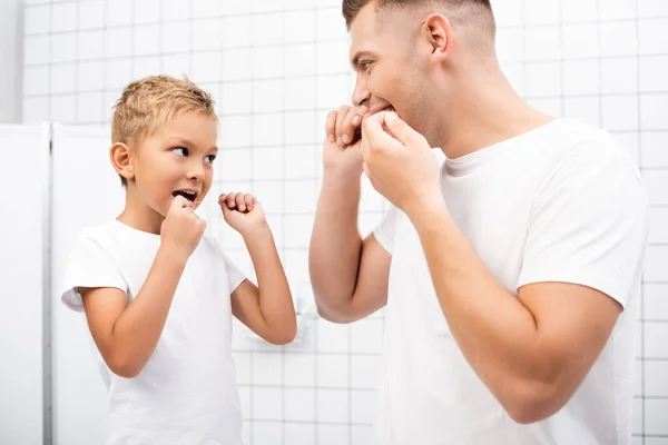 Padre e hijo mirándose mientras limpian los dientes con hilo dental en el baño - foto de stock