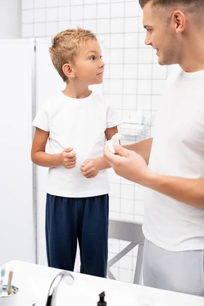 Emocionado niño preescolar mirando a padre con soporte de hilo dental mientras está de pie en la silla en el baño - foto de stock