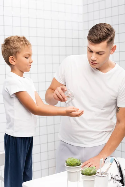 Vater schüttet Rasierwasser auf Hände seines Sohnes, der auf Stuhl neben Waschbecken im Badezimmer steht — Stockfoto