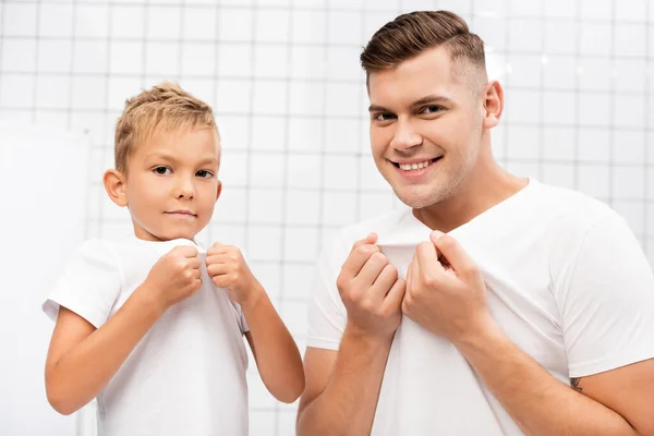 Lächelnder Vater und Sohn ziehen weiße T-Shirts, während sie im Badezimmer in die Kamera schauen — Stockfoto