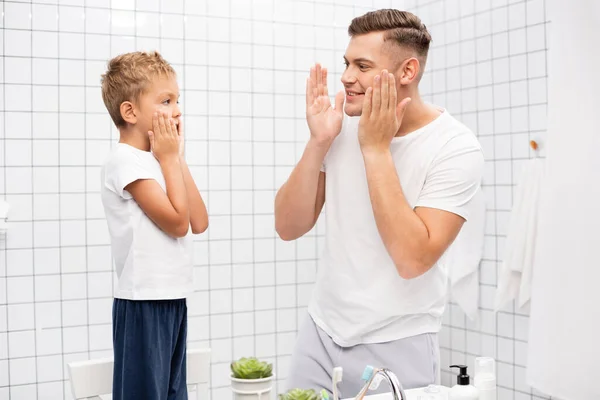 Sorridente padre con le mani vicino guance guardando il figlio applicare lozione da barba mentre in piedi sulla sedia in bagno — Foto stock