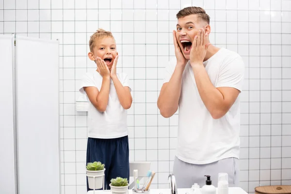Père et fils avec les bouches ouvertes touchant les joues avec les mains, regardant la caméra tout en se tenant près de l'évier dans la salle de bain — Photo de stock
