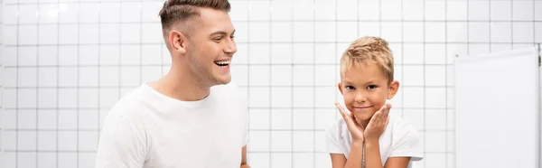 Alegre padre riendo, mientras mira al hijo con las manos cerca de las mejillas en el baño, pancarta - foto de stock