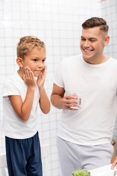 Веселий батько сміється і дивиться на сина, застосовуючи лосьйон для гоління, стоячи на стільці у ванній — стокове фото