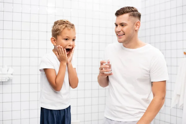 Улыбающийся отец с лосьоном для бритья глядя на шокированного сына с руками на щеках в ванной комнате — стоковое фото