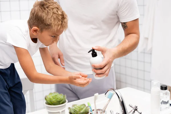 Homme serrant du savon liquide sur la main du garçon penché vers l'avant, tout en se tenant sur une chaise dans la salle de bain — Photo de stock