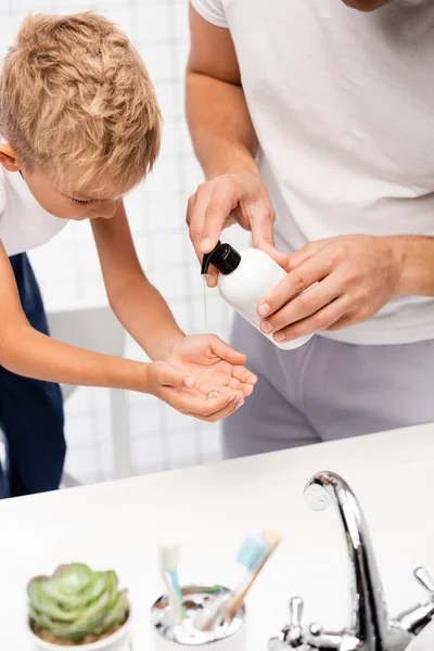 Vater drückt Flüssigseife auf die Hand des Sohnes, der sich nach vorne lehnt, während er im Badezimmer auf einem Stuhl im verschwommenen Vordergrund steht — Stockfoto