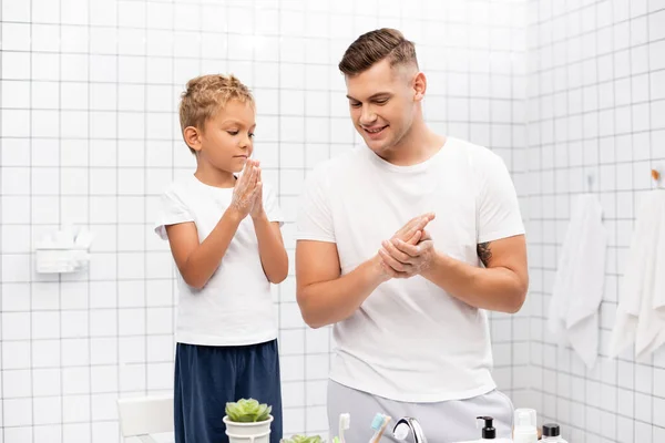 Fils frottant savon entre les mains, tout en se tenant sur la chaise près du père souriant dans la salle de bain — Photo de stock