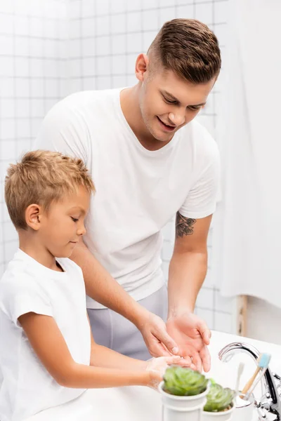 Улыбающиеся отец и сын моют руки, стоя возле раковины в ванной — стоковое фото