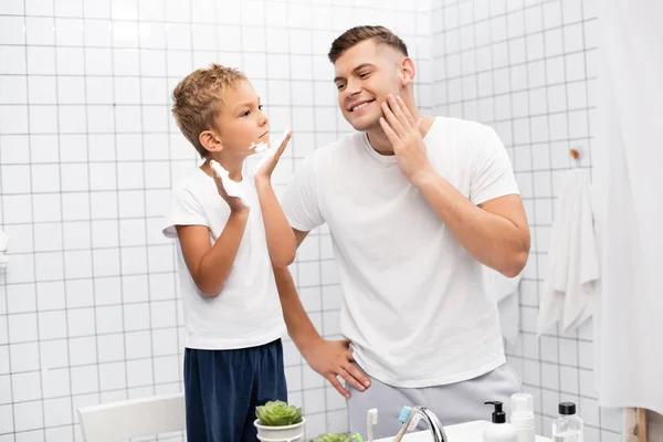 Sorrindo pai com a mão no quadril olhando para o filho aplicando espuma de barbear, enquanto em pé na cadeira no banheiro — Fotografia de Stock