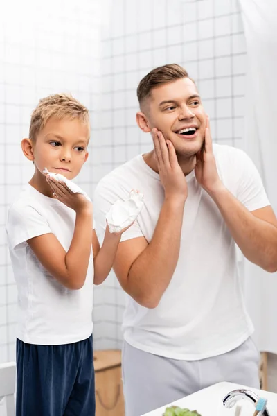 Preschooler figlio applicare schiuma da barba mentre in piedi vicino felice padre toccare le guance in bagno — Foto stock