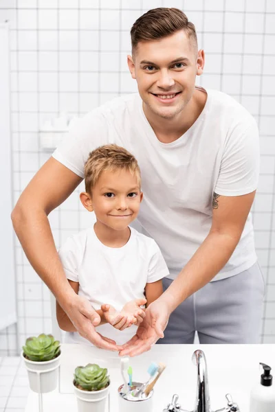 Sonriente padre abrazando al hijo con las manos jabonosas, mientras está de pie detrás y mirando a la cámara en el baño - foto de stock