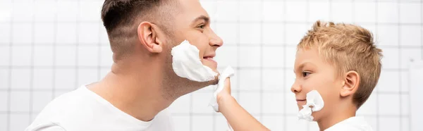 Vista laterale del figlio che applica schiuma da barba sul viso del padre sorridente in bagno, banner — Foto stock