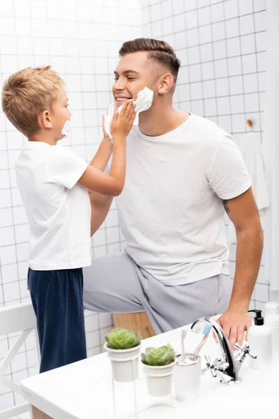 Син наносить піну для гоління на обличчя щасливого батька, стоячи на стільці у ванній — стокове фото