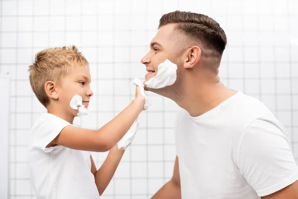 Vue latérale du fils appliquant de la mousse à raser sur le visage du père heureux dans la salle de bain — Photo de stock