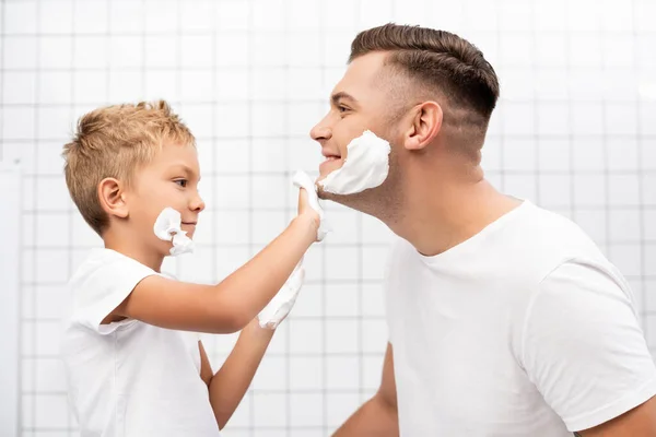Seitenansicht des Sohnes, der Rasierschaum auf das Gesicht des glücklichen Vaters im Badezimmer aufträgt — Stockfoto