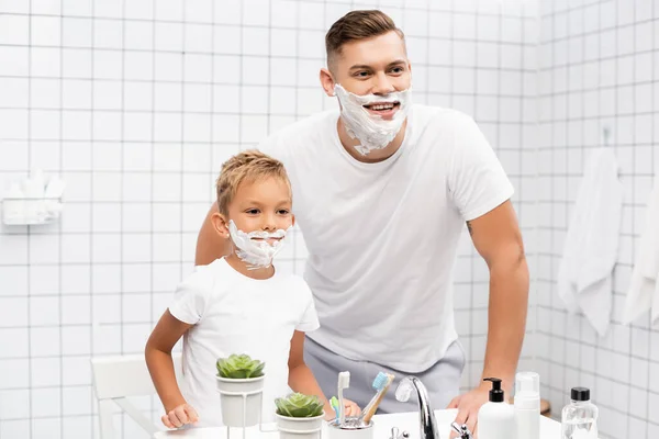 Улыбающиеся отец и сын с пеной для бритья на лицах, стоящих возле раковины в ванной комнате — стоковое фото