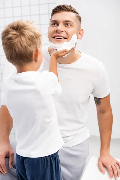 Vista posterior del hijo aplicando espuma de afeitar en la cara del padre sonriente mirando hacia otro lado en el baño - foto de stock
