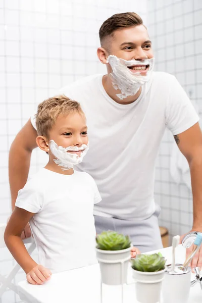 Щасливий син з піною для гоління на обличчі, що стоїть біля усміхненого батька, спираючись на раковину у ванній — стокове фото