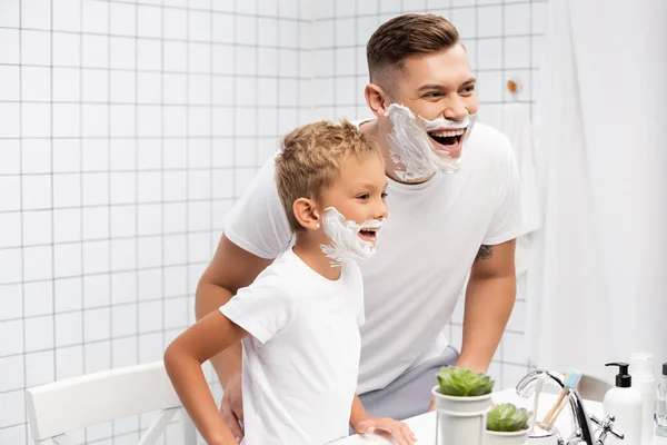 Vater und Sohn mit Rasierschaum auf lachenden Gesichtern, während sie sich im Badezimmer auf Waschbecken mit Toilettenartikeln stützen — Stockfoto
