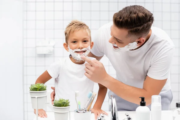 Sonriente padre abrazando y tocando la barbilla del hijo con espuma de afeitar en la cara en el baño - foto de stock