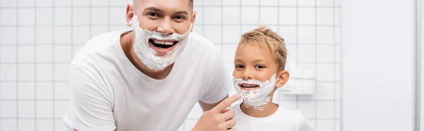 Щасливий батько стоїть біля сина з піною для гоління на обличчі і торкається підборіддя у ванній кімнаті, банер — стокове фото