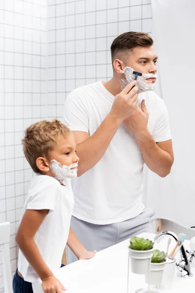 Menino com espuma de barbear no rosto encostado na pia enquanto estava perto do homem usando navalha de segurança no banheiro — Fotografia de Stock