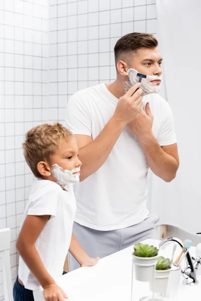 Niño con espuma de afeitar en la cara apoyada en el fregadero, mientras que de pie cerca del hombre utilizando la máquina de afeitar de seguridad en el baño - foto de stock